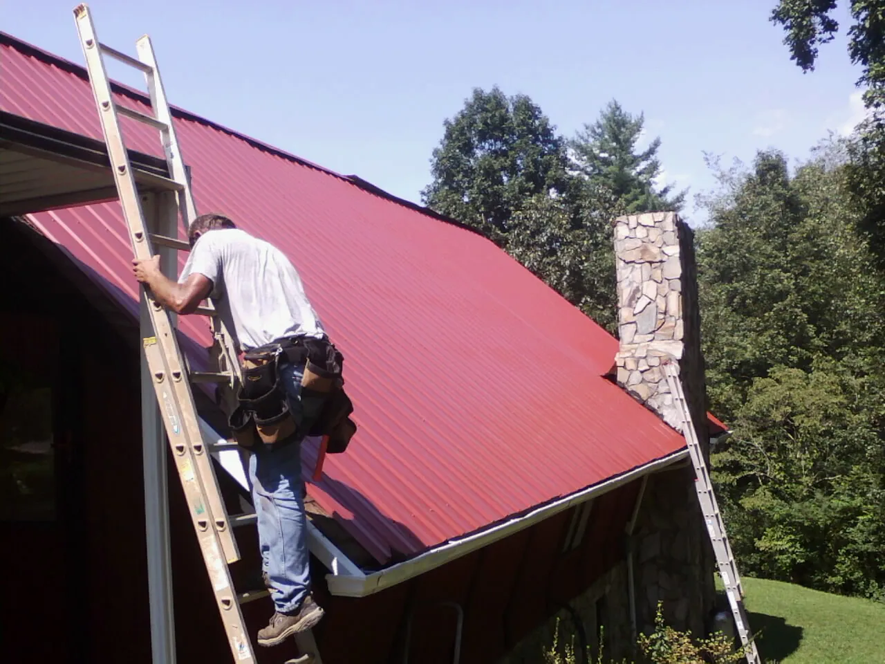 Roofer on a ladder beside a red metal roof and stone chimney — Soffit & Fascia Repair by Roofing Brothers in Ibapah